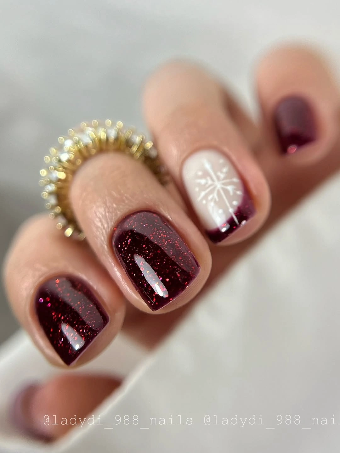 Short burgundy glitter nails with a pale accent and white snowflake design.