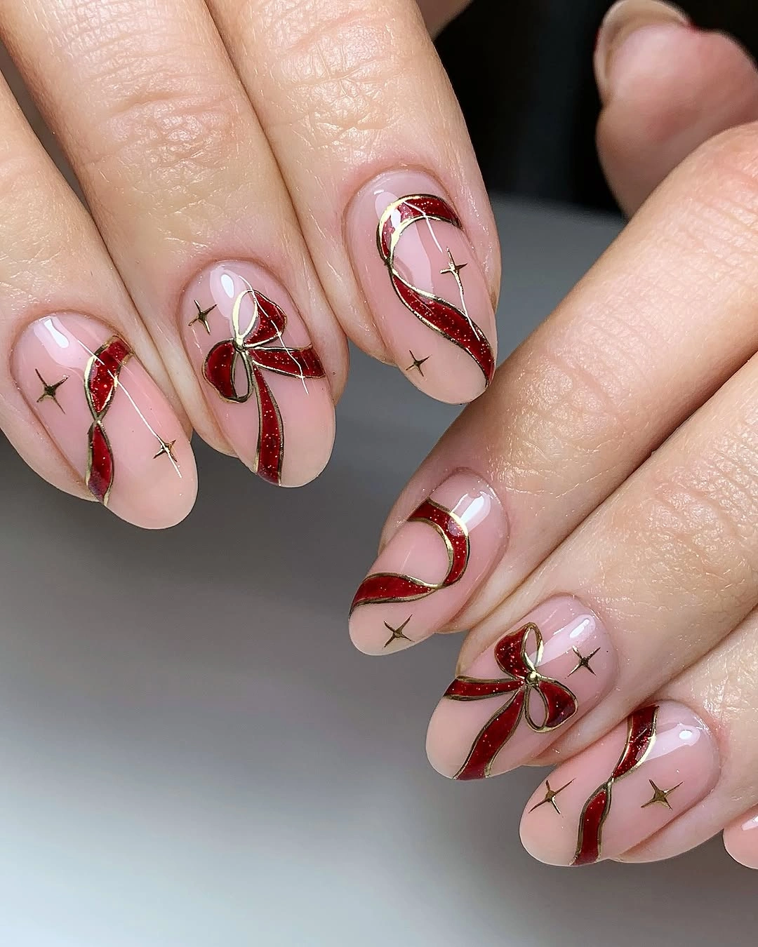 Nude pink almond nails with red glitter ribbons, gold outlines and bows.