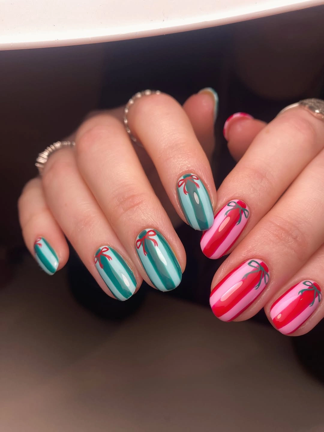 Teal and pink oval nails with contrasting stripes and small festive ribbons.