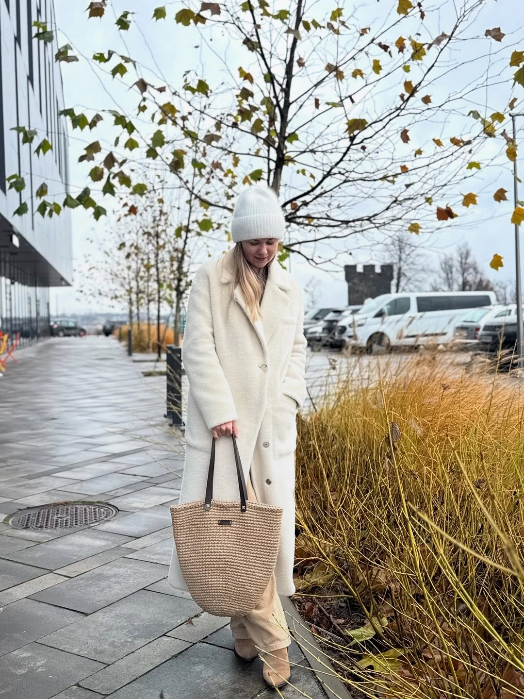 Long cream coat with white knit beanie and tan woven-look tote.