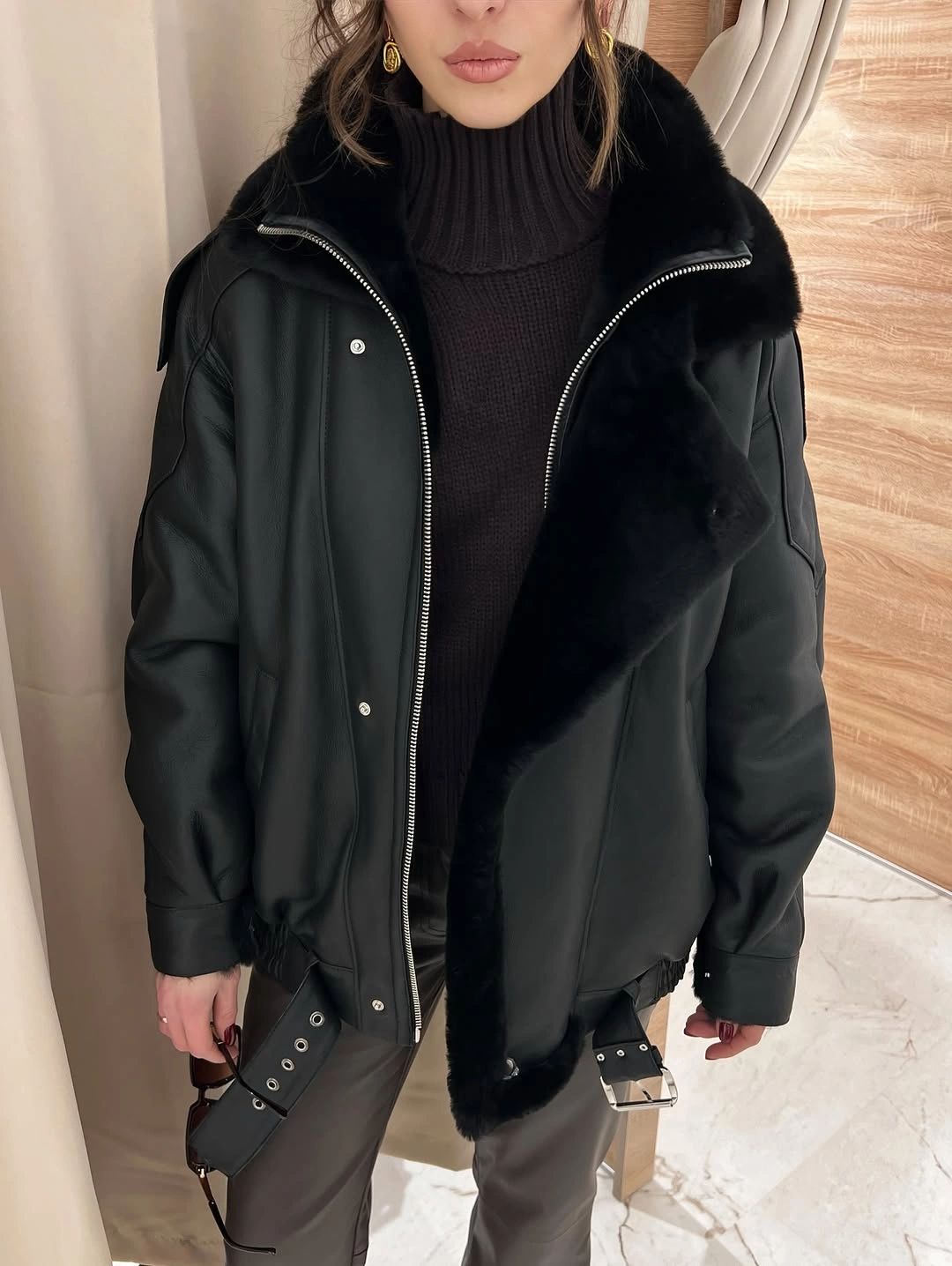 Black leather-look jacket with plush collar, dark turtleneck, and gray trousers.